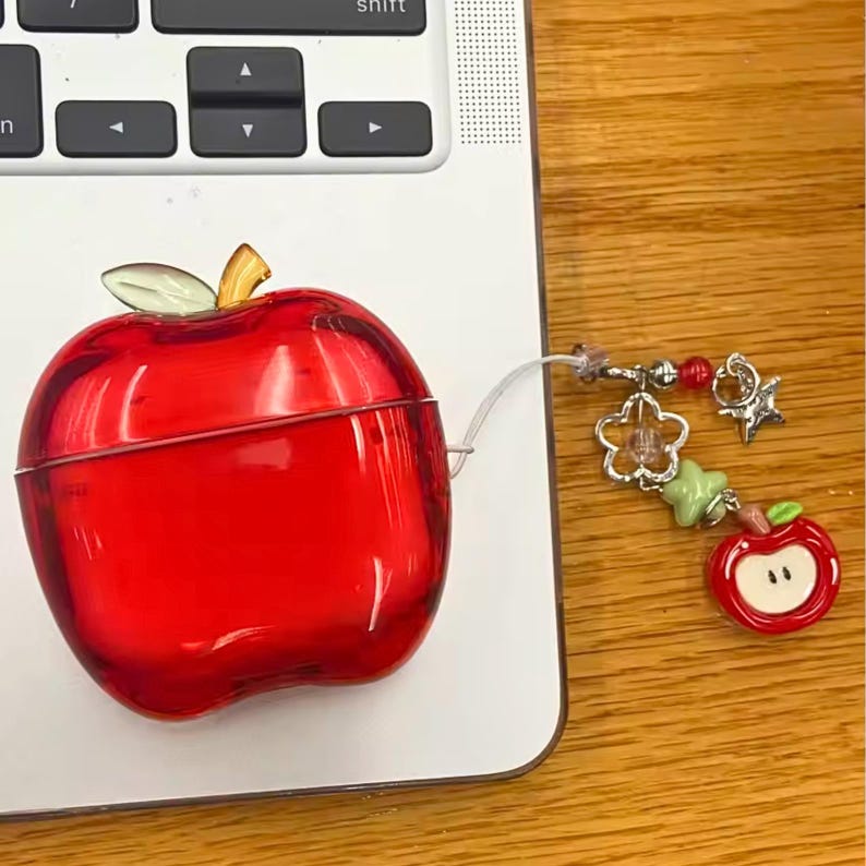 Puede incluir: Una funda roja con forma de manzana para auriculares inal&aacute;mbricos. La funda tiene un acabado transparente y brillante y un tallo y una hoja dorados. Se adjunta un dije con una flor, una estrella y una cuenta de manzana. La funda est&aacute; sobre una superficie blanca.