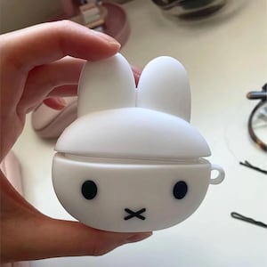 Op de afbeelding: Een witte, konijnvormige hoes voor draadloze oordopjes. De hoes heeft een afgeronde behuizing met twee zwarte stipogen en een zwarte 'x'-mond. De bovenkant van de hoes heeft twee rechtopstaande oren. De hoes wordt in een hand gehouden.