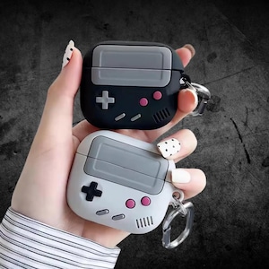 May include: Two AirPod cases in black and white, styled like retro handheld game consoles. They have grey screens, pink buttons, and directional pads. Each case includes a keychain attachment.