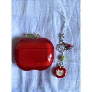 Puede incluir: Un recipiente rojo en forma de manzana con una hoja verde y un tallo dorado, junto a un dije con un colgante de manzana, tr&eacute;boles y dijes en forma de estrella. El recipiente es probablemente un estuche.