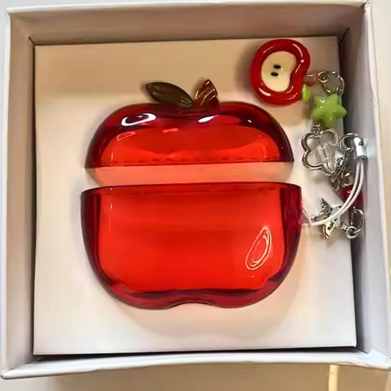 Puede incluir: Una funda roja con forma de manzana, abierta para revelar un interior rosa claro, se encuentra dentro de una caja blanca. Un peque&ntilde;o dije de manzana y un llavero con dijes est&aacute;n adjuntos. La funda est&aacute; hecha de un material brillante y transparente.