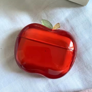 Puede incluir: Un recipiente rojo brillante con forma de manzana con una hoja verde y un tallo dorado. El recipiente est&aacute; hecho de un material transparente que refleja la luz. El dise&ntilde;o es un objeto decorativo.