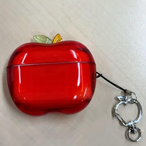 Puede incluir: Una funda roja con forma de manzana para auriculares inal&aacute;mbricos. La funda tiene un detalle de hoja verde y amarilla en la parte superior y un llavero plateado con forma de manzana. La funda est&aacute; hecha de un material transl&uacute;cido.