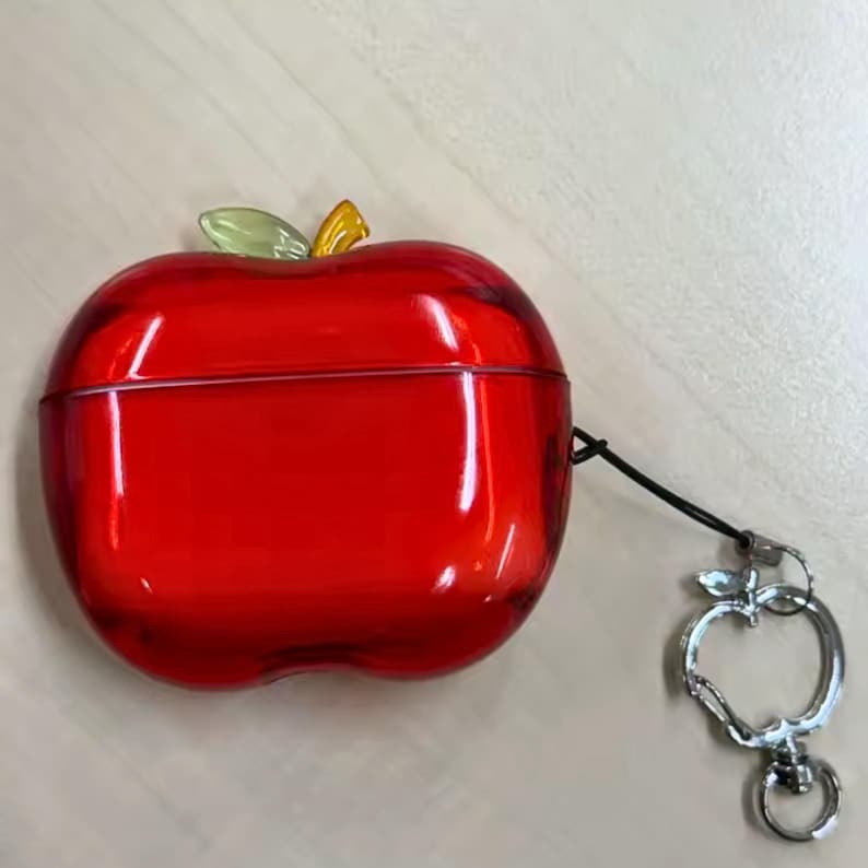 Puede incluir: Una funda roja con forma de manzana para auriculares inal&aacute;mbricos. La funda tiene un detalle de hoja verde y amarilla en la parte superior y un llavero plateado con forma de manzana. La funda est&aacute; hecha de un material transl&uacute;cido.