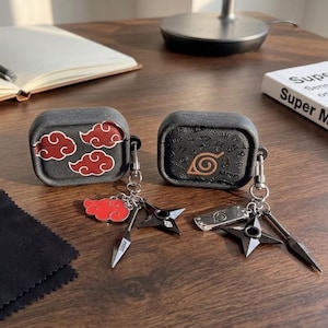 Peut inclure: Deux étuis de protection noirs pour écouteurs sans fil. L'un présente des motifs de nuages rouges, l'autre un symbole de feuille beige et de petites icônes gravées. Les deux incluent des porte-clés métalliques avec des breloques en forme d'étoiles et de lames.