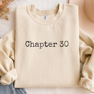 May include: A cream-colored sweatshirt with the text "Chapter 30" in black. The sweatshirt is folded, with the sleeves and bottom hem visible. A neutral-toned hat and a pair of blue jeans are partially visible in the background.