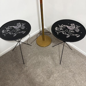 May include: Two round black metal side tables with thin tripod legs, each featuring a white line-art illustration of a dancing couple on the tabletop surface. A tall floor lamp with a brass base and wooden pole stands between the two tables.