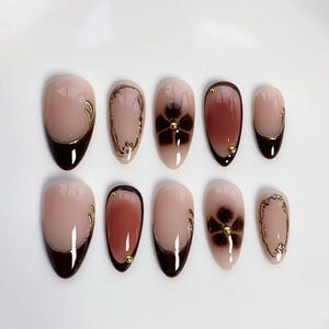 Op de afbeelding: Set van tien amandelvormige valse nagels. De nagels hebben een nude basis met diverse ontwerpen, waaronder donkerbruine tips, bloemaccenten en gouden versieringen. Sommige nagels hebben een donkerbruine gradiënt, andere gouden krullen.