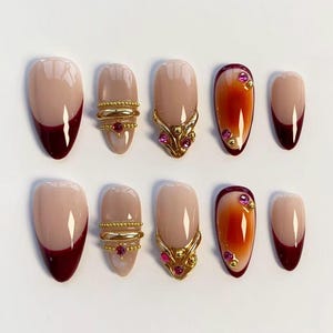 Puede incluir: Un conjunto de diez uñas artificiales con varios diseños. Algunas uñas tienen una base nude con una punta burdeos oscuro. Otras tienen adornos dorados con gemas rosas y un efecto degradado.