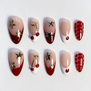 Pode incluir: Um conjunto de dez unhas postiças em forma de amêndoa. As unhas apresentam uma variedade de designs, incluindo estrelas douradas, cerejas e uma ponta francesa vermelha e branca. Uma unha tem um padrão de réptil vermelho.