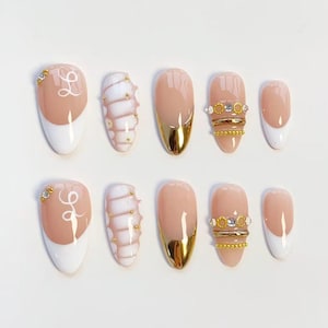 Peut inclure: Un ensemble de dix faux ongles aux designs variés. Certains ongles sont nude avec des accents dorés, d'autres ont des pointes blanches, et certains présentent des ornements dorés et une lettre cursive. Les ongles sont de forme ovale.