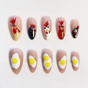 Può includere: Un set di dieci unghie artificiali con una base beige chiaro. La fila superiore presenta disegni di galli in rosso, nero e bianco. La fila inferiore presenta disegni di uova fritte con tuorli gialli e albumi bianchi.