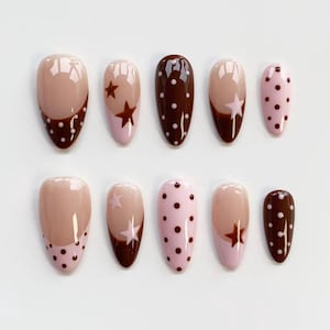 Puede incluir: Un conjunto de diez u&ntilde;as postizas en forma de almendra. Las u&ntilde;as presentan una variedad de dise&ntilde;os, incluyendo marr&oacute;n liso, rosa con lunares marrones y nude con detalles de estrellas y puntos marrones. Las u&ntilde;as est&aacute;n dispuestas sobre una superficie blanca.