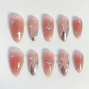 Puede incluir: Un conjunto de diez uñas postizas en forma de almendra. Cada uña presenta un diseño degradado, que transiciona de un rosa claro a un tono melocotón más oscuro. Algunas uñas tienen detalles plateados en forma de estrella, otras gotas metálicas plateadas y pequeños diamantes de imitación.