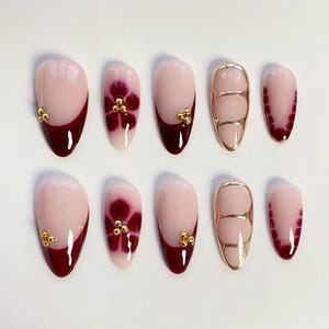 May include: A set of ten artificial nails in various designs. Some nails feature a light pink base with a dark red French tip and gold accents. Others have a floral design or a gold metallic frame. The nails are oval-shaped.