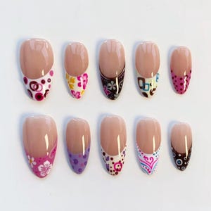 Op de afbeelding: Een set van tien kunstnagels met een nude basis en diverse decoratieve uiteinden. Ontwerpen omvatten bloempatronen, stippen en geometrische vormen in kleuren als roze, paars en bruin. De nagels zijn ovaal van vorm.