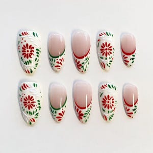 Op de afbeelding: Een set van tien kunstnagels in een French manicure-stijl met een nude basis. De uiteinden zijn versierd met een rood en groen bloemenpatroon, inclusief rode madeliefachtige bloemen en groene bladaccenten op een witte achtergrond.