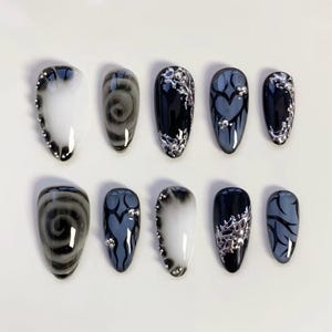 Puede incluir: Un conjunto de diez uñas artificiales con varios diseños. Algunas uñas presentan patrones grises arremolinados, mientras que otras tienen adornos plateados y diseños de corazón azul oscuro y gris. Las uñas son de forma ovalada y están dispuestas sobre una superficie blanca.