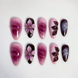 May include: A collection of ten artificial nails in diverse designs. The nails display a gradient of pink and purple shades, with some decorated with 3D floral decorations and others with abstract patterns. The nails are oval-shaped.