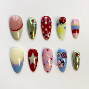 May include: A set of ten artificial nails in diverse designs. The nails showcase colours such as pink, red, green, and blue, with patterns including polka dots, stars, and swirls. Some nails are decorated with small embellishments.