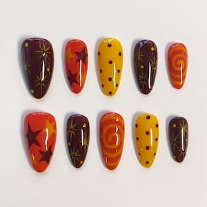 May include: A set of ten almond-shaped press-on nails featuring a warm colour palette of deep burgundy, bright orange, and golden yellow. The designs include dark starburst patterns, solid orange stars, vertical rows of dark dots, and orange spirals.