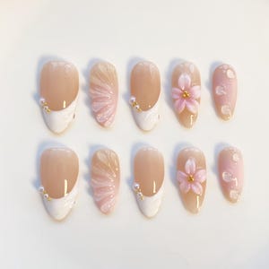 May include: A set of ten artificial nails in various designs. The nails feature a nude base with white tips, pink floral accents, and pearl embellishments. Some nails have a fan-like design in pink.