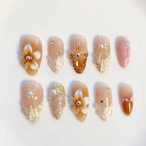 May include: A set of ten artificial nails in various designs. The nails feature a range of colors, including nude, pink, and gold, with embellishments such as flowers, rhinestones, and glitter. Some nails have a textured, layered appearance.