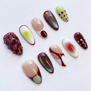 Peut inclure: Une s&eacute;lection de dix faux ongles aux designs vari&eacute;s. Les ongles pr&eacute;sentent des couleurs comme le bordeaux, le beige, le vert et le blanc, avec des motifs incluant des pois, des paillettes et des formes abstraites. Les ongles sont dispos&eacute;s sur une surface blanche.
