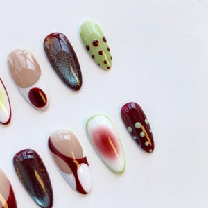 Peut inclure: Une collection de faux ongles en forme d'amande avec divers motifs. Les ongles pr&eacute;sentent des couleurs comme le beige, le bordeaux, le vert et le blanc, avec des motifs tels que des pois, des blocs de couleurs et des effets paillet&eacute;s. Les ongles sont dispos&eacute;s sur une surface blanche.