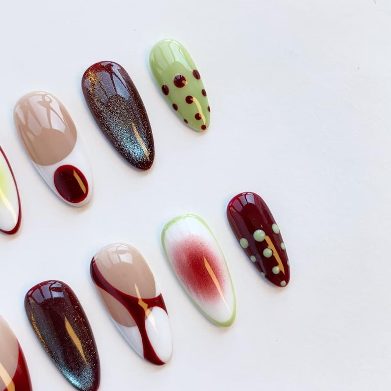 Peut inclure: Une collection de faux ongles en forme d'amande avec divers motifs. Les ongles pr&eacute;sentent des couleurs comme le beige, le bordeaux, le vert et le blanc, avec des motifs tels que des pois, des blocs de couleurs et des effets paillet&eacute;s. Les ongles sont dispos&eacute;s sur une surface blanche.