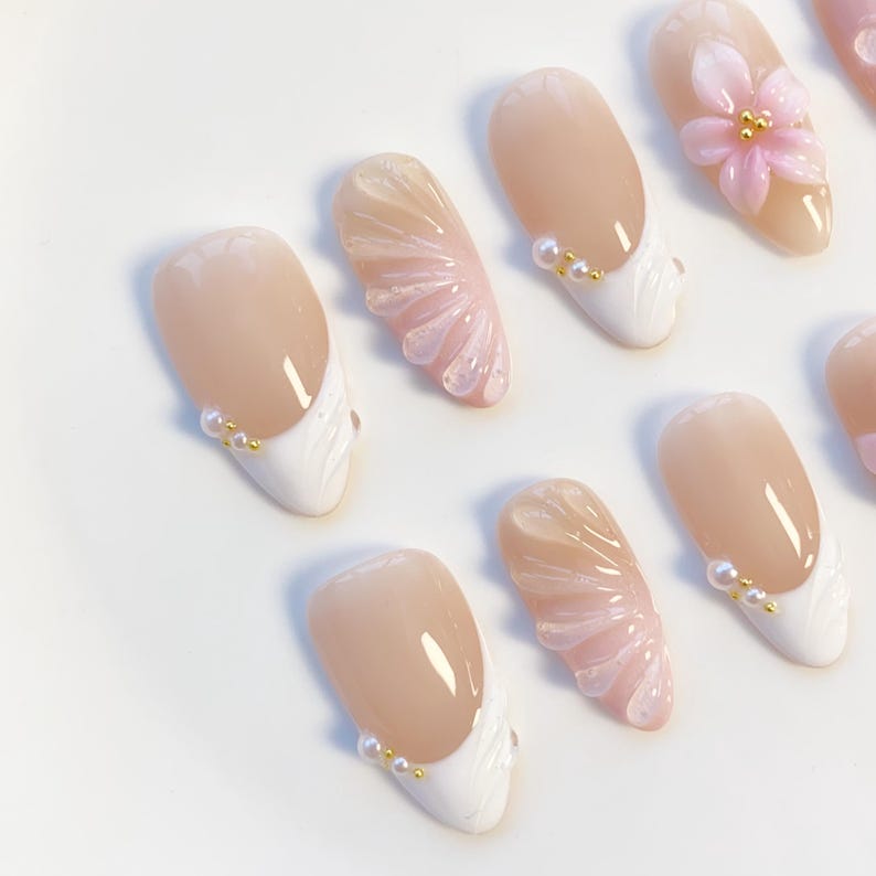 Pu&ograve; includere: Un set di unghie finte con un design rosa chiaro e bianco. Alcune unghie presentano una punta francese bianca con dettagli di perle, mentre altre hanno un fiore 3D o un design a forma di ventaglio. Finitura lucida.