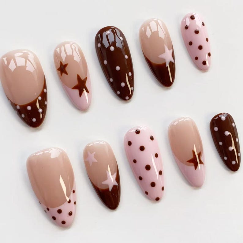 Puede incluir: Un conjunto de diez u&ntilde;as artificiales con varios dise&ntilde;os. Las u&ntilde;as presentan una combinaci&oacute;n de colores rosa claro, marr&oacute;n y nude. Algunas u&ntilde;as tienen dise&ntilde;os de estrellas y lunares, mientras que otras tienen un dise&ntilde;o de punta francesa con lunares.