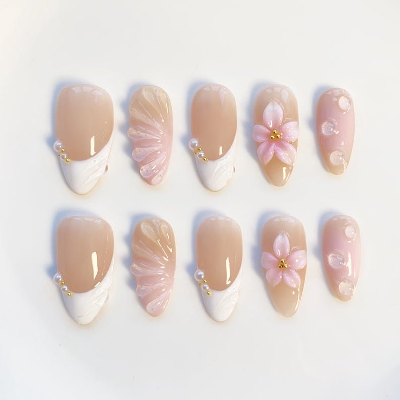 Pu&ograve; includere: Set di dieci unghie finte in vari design. Alcune unghie presentano una base nude con una punta francese bianca e dettagli di perline dorate. Altre hanno fiori rosa e abbellimenti di perle. Le unghie sono di forma ovale.