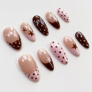 Puede incluir: Una colecci&oacute;n de u&ntilde;as postizas en forma de almendra con varios dise&ntilde;os. Algunas u&ntilde;as son de color rosa liso, otras tienen una base marr&oacute;n con un dise&ntilde;o de estrella, y otras tienen una base rosa con lunares marrones. Las u&ntilde;as est&aacute;n dispuestas sobre una superficie blanca.