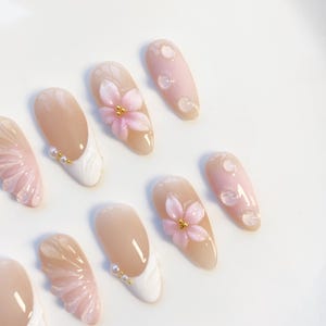 Pu&ograve; includere: Una collezione di unghie finte in vari design. Alcune unghie presentano una base nude con punte bianche e dettagli di perle. Altre presentano decorazioni floreali rosa e dettagli di goccioline trasparenti, il tutto su sfondo bianco.