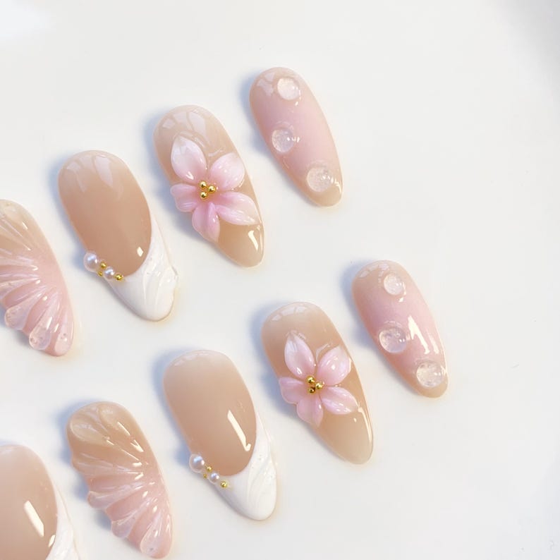 Pu&ograve; includere: Una collezione di unghie finte in vari design. Alcune unghie presentano una base nude con punte bianche e dettagli di perle. Altre presentano decorazioni floreali rosa e dettagli di goccioline trasparenti, il tutto su sfondo bianco.