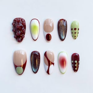 Peut inclure: Une collection de dix faux ongles aux designs vari&eacute;s. Les ongles pr&eacute;sentent des couleurs bordeaux, beige, vert et blanc, avec des motifs tels que des pois, des tourbillons et des effets de bulles textur&eacute;es. Les ongles sont dispos&eacute;s sur une surface blanche.