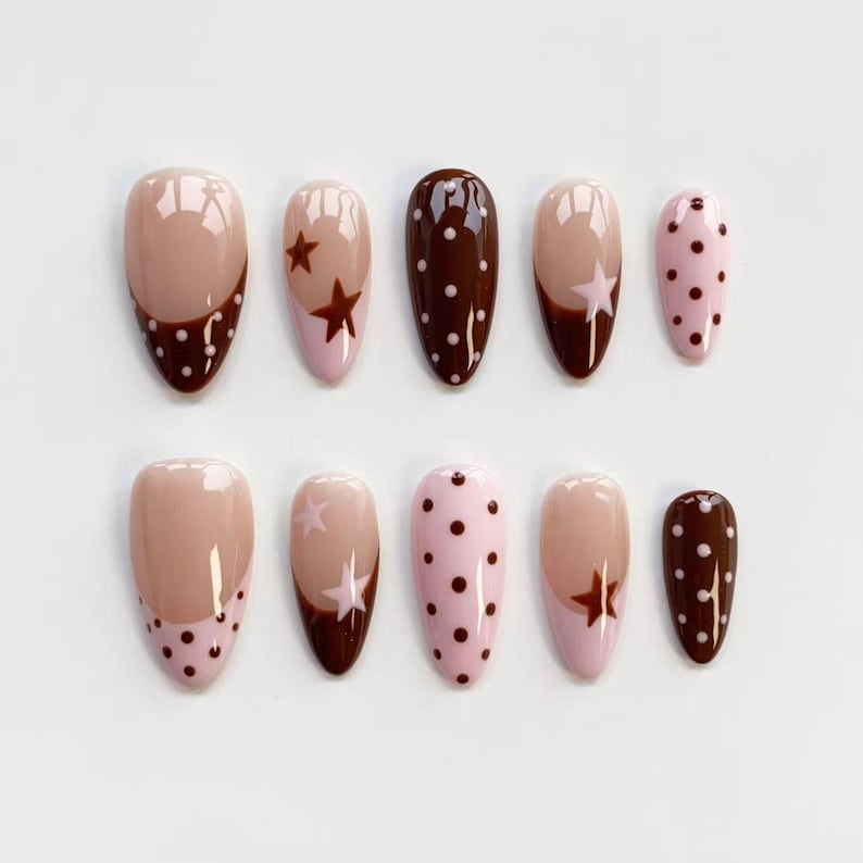 Puede incluir: Un conjunto de diez u&ntilde;as postizas en forma de almendra. Las u&ntilde;as presentan una variedad de dise&ntilde;os, incluyendo marr&oacute;n liso, rosa con lunares marrones y nude con detalles de estrellas y puntos marrones. Las u&ntilde;as est&aacute;n dispuestas sobre una superficie blanca.