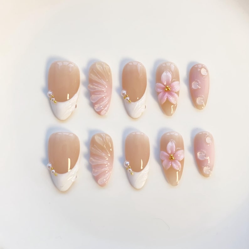 May include: A set of ten artificial nails. The nails have a nude base with white tips, pink floral accents, and pearl embellishments. Some nails feature a fan-like design in pink. The nails are oval-shaped.