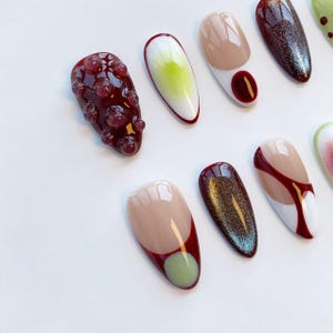 Peut inclure: Une collection d'ongles artificiels avec divers motifs. Certains ongles sont bordeaux avec des embellissements en forme de bulles, d'autres pr&eacute;sentent des d&eacute;grad&eacute;s, des paillettes et des motifs abstraits dans des tons beige, rouge et vert. Les ongles sont dispos&eacute;s sur une surface blanche.