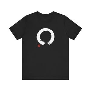 May include: Black t-shirt featuring a large white brushstroke circle design. Below the circle is a small red square with a symbol. The shirt is a short-sleeved crew neck.