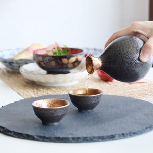 以下が含まれることがあります： 黒いセラミック製の酒器セット。金色の内側があり、2つの小さなカップに液体を注ぐカラフが含まれています。セットは、濃い灰色の丸いスレートサービングトレイにあります。他のボウルが背景にあります。