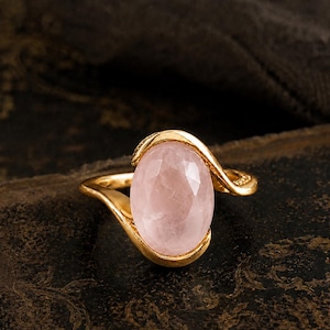 Op de afbeelding: Een gouden ring met een grote, ovaal geslepen rozenkwarts edelsteen. De ring heeft een uniek, vloeiend ontwerp. De rozenkwarts heeft een zachte roze tint, tegen een donkere achtergrond.