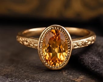 Puede incluir: Un anillo de oro con una piedra preciosa de color ámbar de corte ovalado engastada en un bisel con cuentas. La banda está hecha de oro amarillo y muestra intrincados patrones florales grabados en los lados, dando textura al metal.