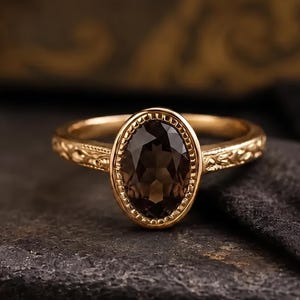 Pode incluir: Um anel de ouro com uma pedra preciosa castanho-escura de corte oval engastada numa moldura com contas. A banda é feita de ouro amarelo e apresenta um padrão floral intrincado em relevo nas laterais, estendendo-se a partir do engaste.