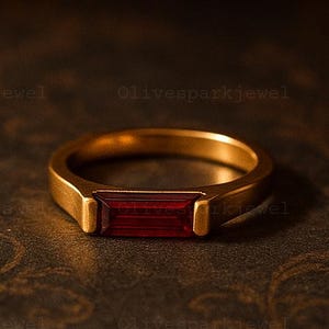 Op de afbeelding: Een goudkleurige ring met een rechthoekige, dieprode edelsteen in het midden. De ring heeft een eenvoudig, elegant ontwerp met een gladde band en een gepolijste afwerking. De edelsteen is ongeveer 1,3 cm lang.