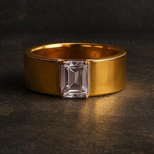 May include: A gold-colored ring with a rectangular, clear gemstone set in the center. The ring has a wide band and a polished finish, set against a dark, textured background. The ring is a piece of jewelry.
