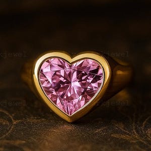 Könnte beinhalten: Ein goldener Ring mit einem herzförmigen rosa Edelstein. Der Edelstein ist in einer goldenen Lünette gefasst, und der Ring hat einen glatten Band. Der Ring wird auf einer dunklen, gemusterten Oberfläche präsentiert.