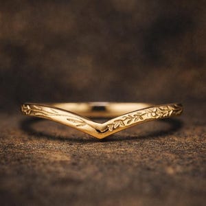 May include: A gold-colored ring with a V-shaped design. The ring features delicate floral and leaf patterns along the band. The ring is set against a dark, textured background, highlighting its intricate details.