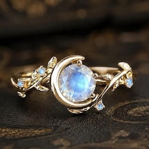 Può includere: Un anello in oro con una pietra di luna rotonda centrale parzialmente incorniciata da una forma a mezzaluna. La fascia è progettata come una vite con piccole foglie e minuscole pietre d'accento incastonate lungo i lati.